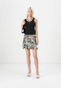 Sort tanktop med en logo, parret med en grå camo plisseret mini-skjørt. Modellen bærer hvide slidesandaler og holder en sort håndtaske.