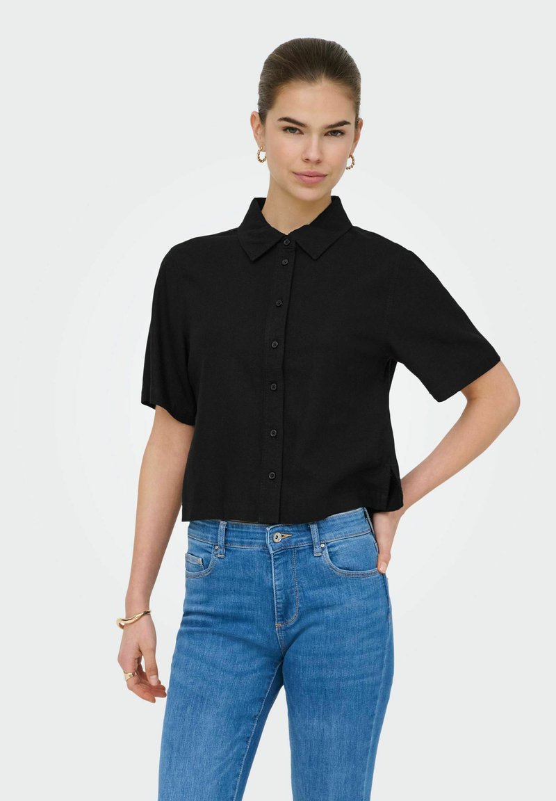 Femme aux cheveux attachés en chignon portant des boucles d'oreilles créoles en or et un bracelet, une chemise noire à manches courtes avec boutons, et un jean bleu taille haute.