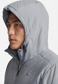 Gray hooded jacket with a smooth, lightweight texture, featuring a zippered front and adjustable drawstring hood. Includes a logo accent.
