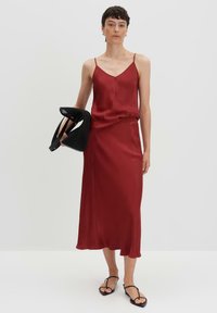 Bordeaux satijnen slipjurk met spaghettibandjes, vloeiende silhoeet en subtiele glans; gecombineerd met een zwarte gaas handtas en minimalistische sandalen.