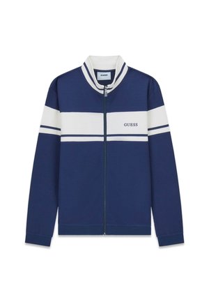 Veste zippée bleu marine avec des rayures horizontales blanches et le logo "GUESS" sur la poitrine, poignets et col côtelés avec des accents blancs et bleus.