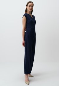 Une combinaison bleu marine en tissu texturé, avec des manches courtes, un col boutonné et des jambes larges, associée à des escarpins beiges à bout pointu.