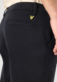Donkerblauwe broek van soepel materiaal heeft een achterzak met een gele geborduurde logo, wat de schone uitstraling en de getailleerde vorm benadrukt.