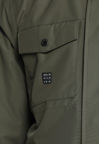 Olivově zelená bunda s hladkou strukturou, vybavená náprsní kapsou uzavřenou černým patentním knoflíkem a značkovou etiketou s nápisem "Quiksilver".