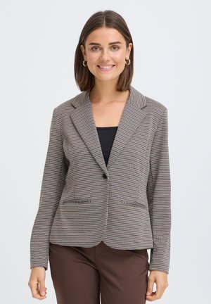 Femme souriante aux cheveux bruns mi-longs, portant des boucles d’oreilles créoles dorées, un blazer à carreaux sur un haut noir et un pantalon marron, debout devant un fond blanc.