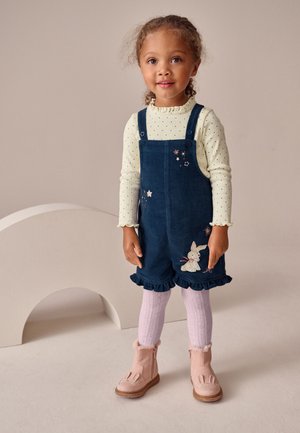 Jong meisje met een crèmekleurige polkadot longsleeve top en marineblauwe tuinbroekshorts met konijnenborduursel, roze panty's en roze laarsjes.