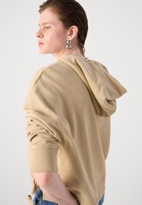 Beige hettegenser med snøring og ribbede mansjetter, med en avslappet passform og rynkedesign bak. Brukt med blå jeans.