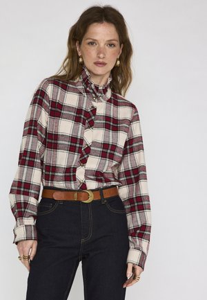 Donna che indossa una camicia a quadri rossa e beige a maniche lunghe con colletto arricciato, cintura marrone, jeans scuri, orecchini a cerchio dorati e anelli alle dita.