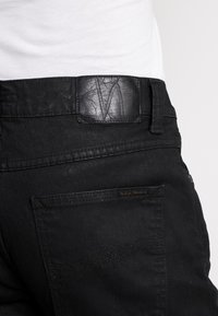 Los jeans de mezclilla negra presentan una cinturilla de cuero negro texturizado con un logo embosado, detalles en los bolsillos traseros y patrones de costura sutiles.