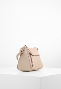 Borsa a mano in pelle beige con forma triangolare, patta superiore e hardware dorato. Presenta una tasca con zip e una tasca frontale. Texture liscia, design minimalista.