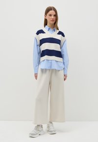 Maglione senza maniche a righe blu e bianche sopra una camicia azzurra, abbinato a pantaloni a gamba larga color crema e sneakers bianche robuste.