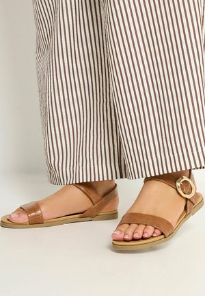 Voeten met bruine platte sandalen met een gouden gesp, gecombineerd met witte broeken met verticale bruine strepen.