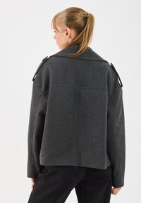 Donna con coda di cavallo bionda che indossa una giacca di lana grigia scura con spalline, girata di spalle su uno sfondo bianco semplice.