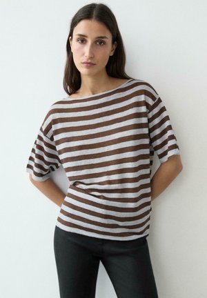 Kobieta o długich ciemnych włosach, ubrana w luźną bluzkę w brązowo-szare paski z krótkim rękawem i czarne spodnie, stoi na tle białej ściany.