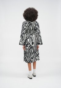 Asmuo su garbanotais plaukais, dėvintis ilgą juodai baltos abstrakčios raštų suknelę, baltas kojines ir baltus batus, stovintis priešais vientisą foną.