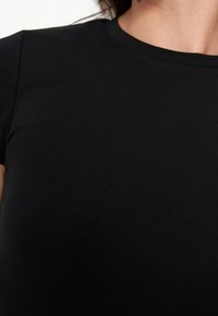 Gros plan d'une personne portant un t-shirt noir uni à manches courtes et col rond sur fond blanc.