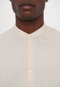 Camicia button-up beige chiaro con colletto alla band, sottili righe verticali e piccoli bottoni bianchi lungo la patta frontale.