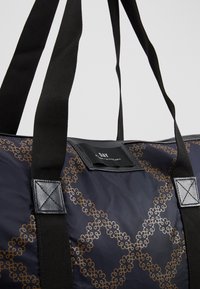 Sac cabas bleu marine avec motif chaîne florale marron entrelacée et anses en tissu noir avec renforcements en cuir cousus.