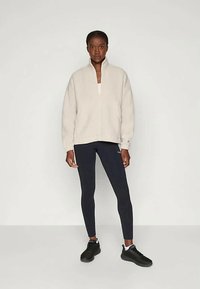 Γυναίκα στέκεται σε απλό φόντο φορώντας ένα κρεμ χρώματος φλις με φερμουάρ έως το τέταρτο, μαύρες κολάν και μαύρα αθλητικά παπούτσια.