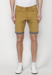 Rumene jeans kratke hlače s zavihanimi robovi, zasnova s petimi žepi, nošene nad kolenom, v kombinaciji z belimi športnimi čevlji in belo majico.
