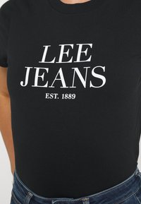 Camiseta negra de algodón, con grandes letras blancas "LEE JEANS" y "EST. 1889" en el frente. Mangas cortas, cuello redondo.