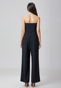 Jumpsuit noir sans bretelles avec un corsage ajusté et un pantalon large, présentant des lignes épurées et une texture douce. Porté avec des talons accentués d'orange.