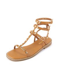 Tan gladiatorsandal med nitar på remmarna och spänne runt vristen. Platt sula, slät lädersula och dekorativa guldnitar längs kanterna.