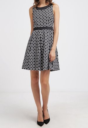 Femme portant une robe sans manches à motifs géométriques noirs et blancs, avec une taille cintrée et une jupe évasée, associée à des escarpins noirs à bouts pointus.