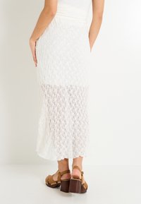 Jupe midi en dentelle blanche, présentant un design texturé avec un panneau inférieur transparent. Associée à des sandales à plateforme marron avec des clous.