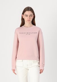 Růžová mikina s žebrovanými manžetami a dolním lemem, se tmavým logem "TOMMY HILFIGER EST. 1985" na hrudi. Nosit s bílými kalhotami.