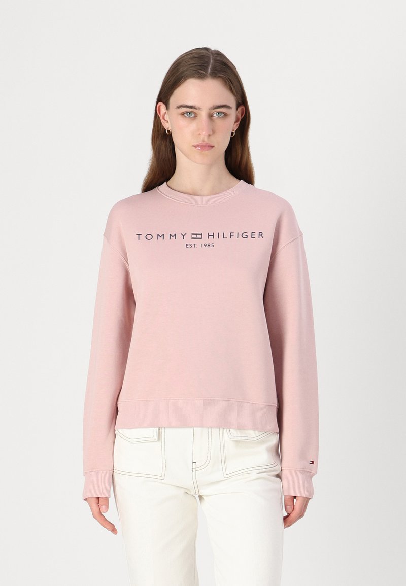 Růžová mikina s žebrovanými manžetami a dolním lemem, se tmavým logem "TOMMY HILFIGER EST. 1985" na hrudi. Nosit s bílými kalhotami.