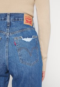 Vue arrière d'une personne portant un jean Levi's bleu avec une poche arrière droite déchirée et un haut à manches longues côtelé beige sur fond blanc.