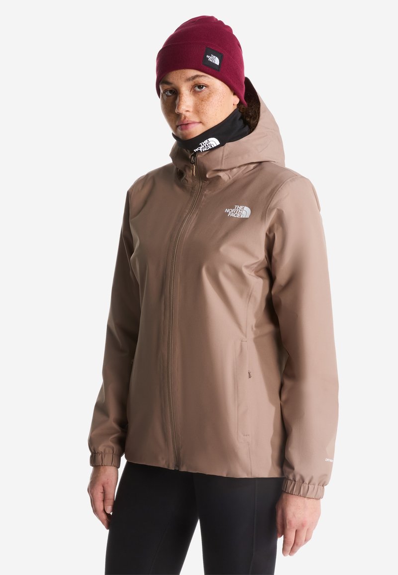Personne portant une veste d'extérieur beige avec une capuche, un cache-cou noir, un bonnet bordeaux et un pantalon noir sur un fond uni.