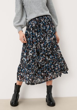 Vrouw die een lange zwarte rok draagt met abstracte blauwe, witte en roze patronen, gecombineerd met zwarte enkellaarsjes en een grijze trui.
