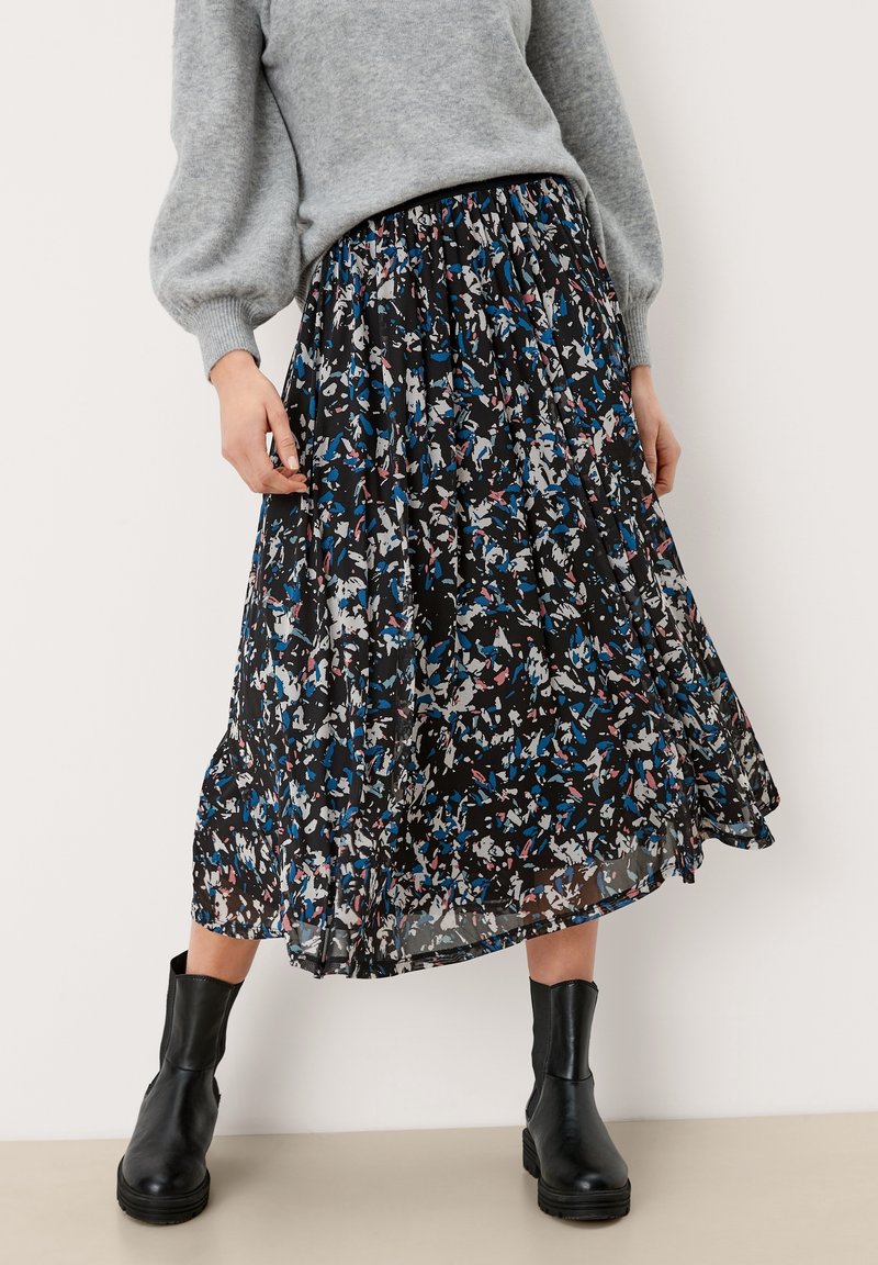 Femme portant une longue jupe noire avec des motifs abstraits bleus, blancs et roses, assortie à des bottines noires et un pull gris.