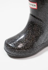 Bota de goma negra con brillo y suela texturizada, que presenta una etiqueta roja en contraste mostrando el nombre de la marca cerca de la parte superior.