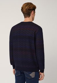 Maglione blu navy con motivo a zigzag, polsini e orlo a costine, lavorazione a maglia texture, cuciture visibili. Indossato da una persona vista da dietro.