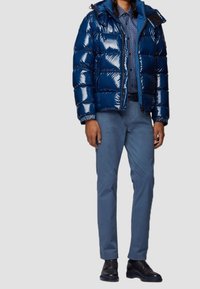 Blå pufferjacka med en glänsande yta, vadderad design, huva och framzipper, i kombination med ljusblå byxor och svarta skor.