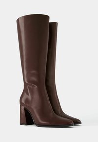 Wysokie ciemnobrązowe skórzane kozaki z ostrymi noskami i grubymi obcasami, stojące obok siebie na jednolitym jasnym tle.