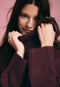 Burgundska turtleneck pulover z rebrastimi manšetami. Model nosi velikansko kovinsko cvetno prstan. Ozadje je mehko roza.