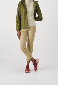 Vihreä nailoninen sadetakki, beige fleece-paita, beiget housut ja punaiset urheilukengät, joissa on vaaleanpunaisia yksityiskohtia. Vetoketjullinen ja kevyt muotoilu.