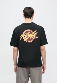 Fekete pamut póló élénk színes grafikával a hátoldalán, amelyen az "Air Jordan Flight" felirat és vízszintes vonalak láthatók, rövid ujjú.