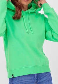 Donna che regola un felpa verde brillante con stringhe, indossa jeans blu, tagliati al collo e in vita su uno sfondo chiaro e semplice.