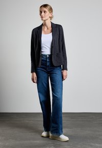 Svart rutig kavaj, vit linne, högmidjade blå jeans och vita sneakers, står på ett grått golv mot en ljus bakgrund.