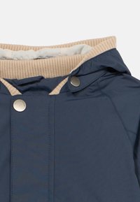 Mornarsko moder plašč z bež rebrastim ovratnikom, opremljen z zaponkami na pritrditev in gladko teksturo. Dizajn vključuje kapuco in vidne šive.