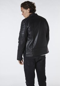 Chaqueta de cuero negra con detalles acanalados en los hombros, textura suave y diseño ajustado. Llevada sobre jeans oscuros, vista desde la parte de atrás.