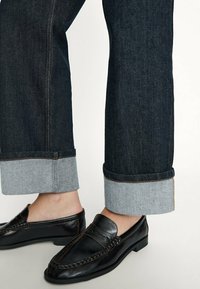 Mörkblå denimjeans med uppvikta fållar; fållarna har en ljusblå accent. Kombinationen med svarta lädersneakers visar detaljerad sömnad.