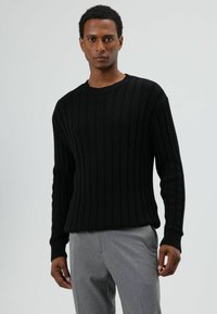 Pull noir en maille côtelée avec un col rond et des manches longues, associé à un pantalon gris clair. La texture semble douce et ajustée.