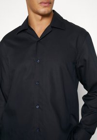 Homme portant une chemise noire à manches longues avec boutons et col décontracté, bras gauche plié visible sur fond blanc.