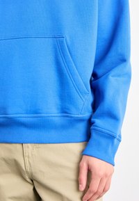Personne portant un sweat-shirt bleu vif avec une poche avant et un pantalon beige, main détendue le long du corps sur un fond uni.